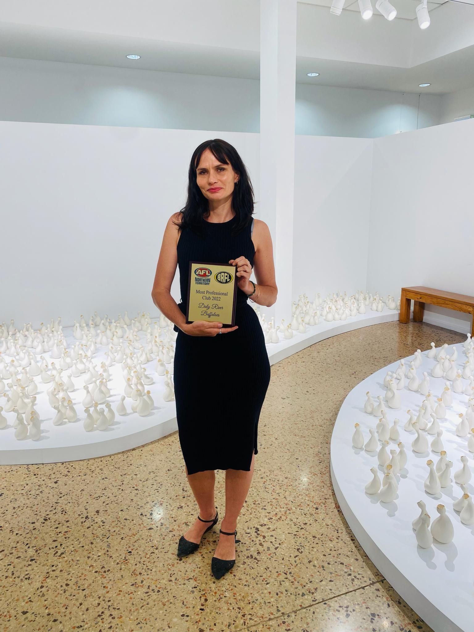 Woman in black dress holds a book, standing in a gallery with white sculptures on the floor.