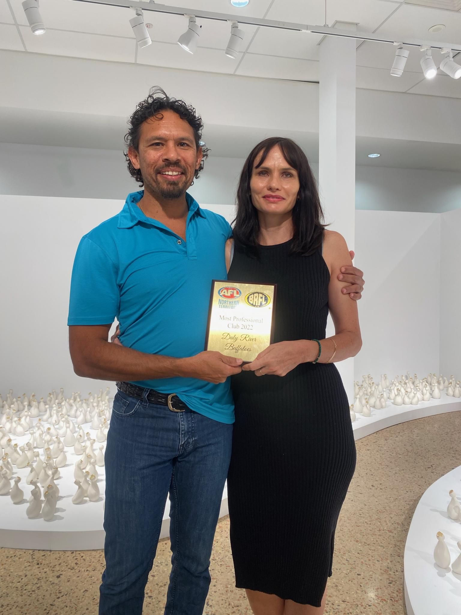 Man and woman smiling, holding award in art gallery with white sculptures.