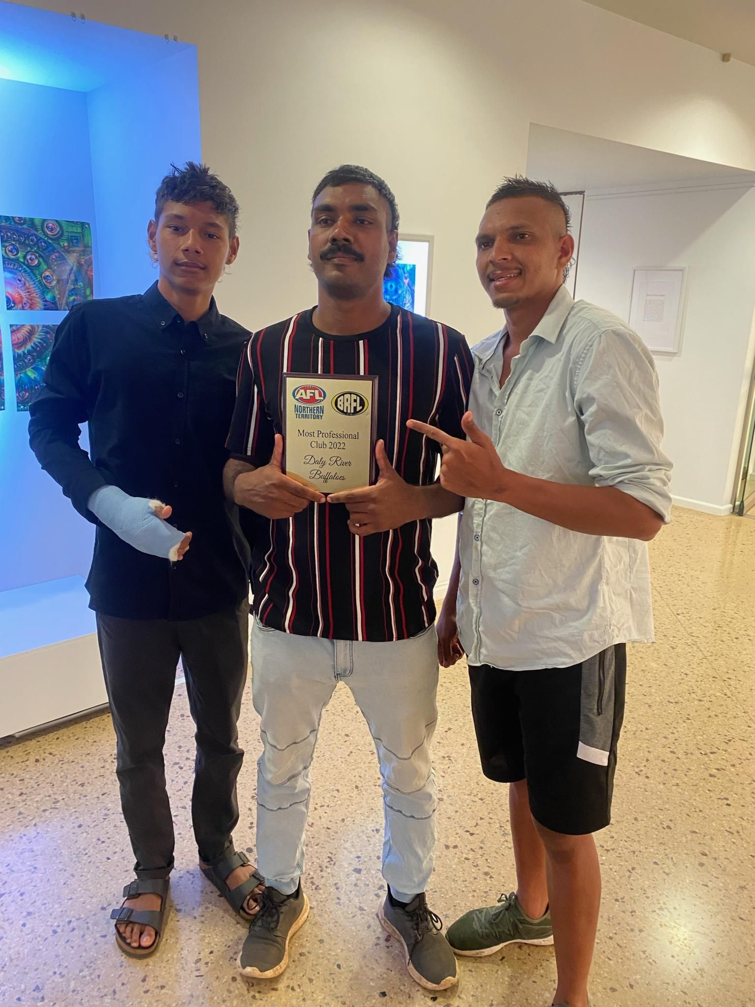 Three men in a brightly lit gallery. Man in the center holds award; others point.
