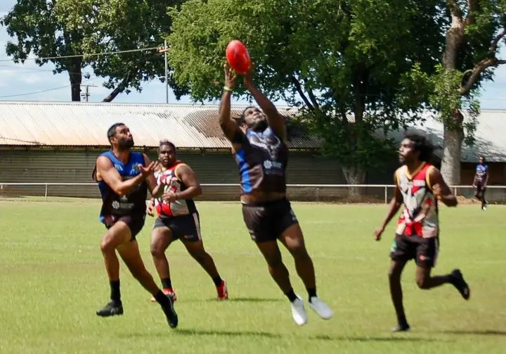 Men playing Australian rules football, one jumping to catch the ball on a green field.