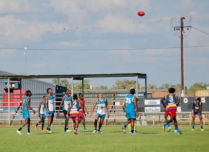 Australian rules football game. Players in blue and maroon uniforms on a green field, ball in the air, sunny day.