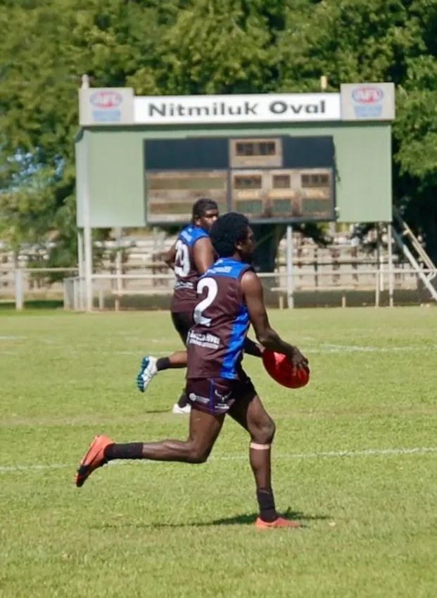 A football player in brown and blue jersey, number 2, runs with the ball on Nitmiluk Oval.