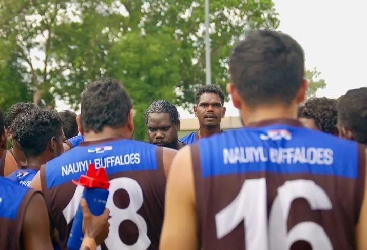 A group of Indigenous Australian Rules footballers wearing brown, blue, and white uniforms huddle together outdoors.