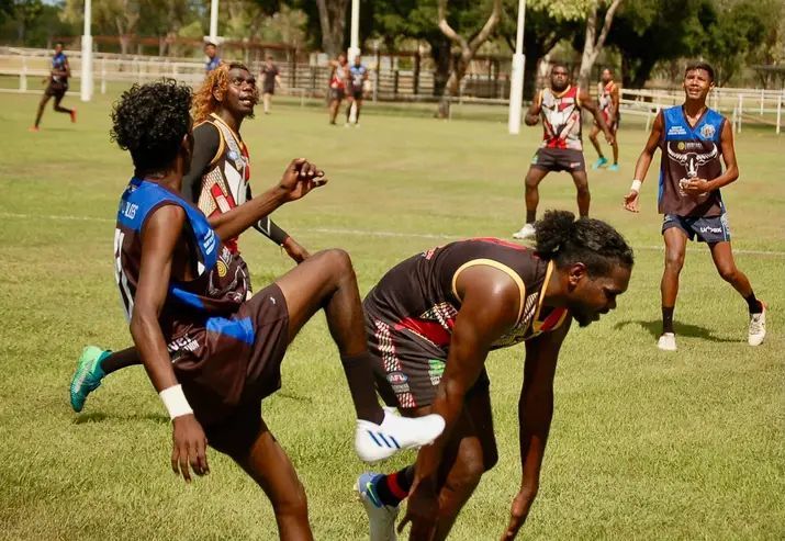 A group of young men playing Australian rules football on a green field in the sun.