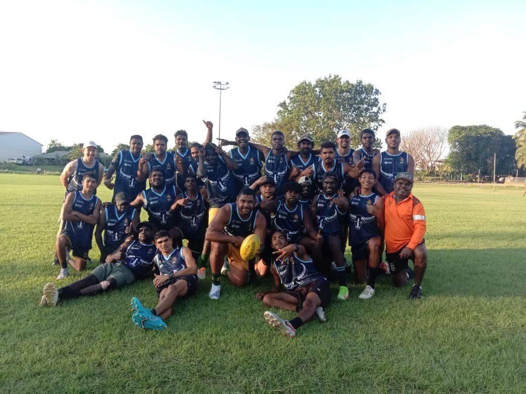 A team of men in blue jerseys and shorts poses on a grassy field, holding an oval ball.