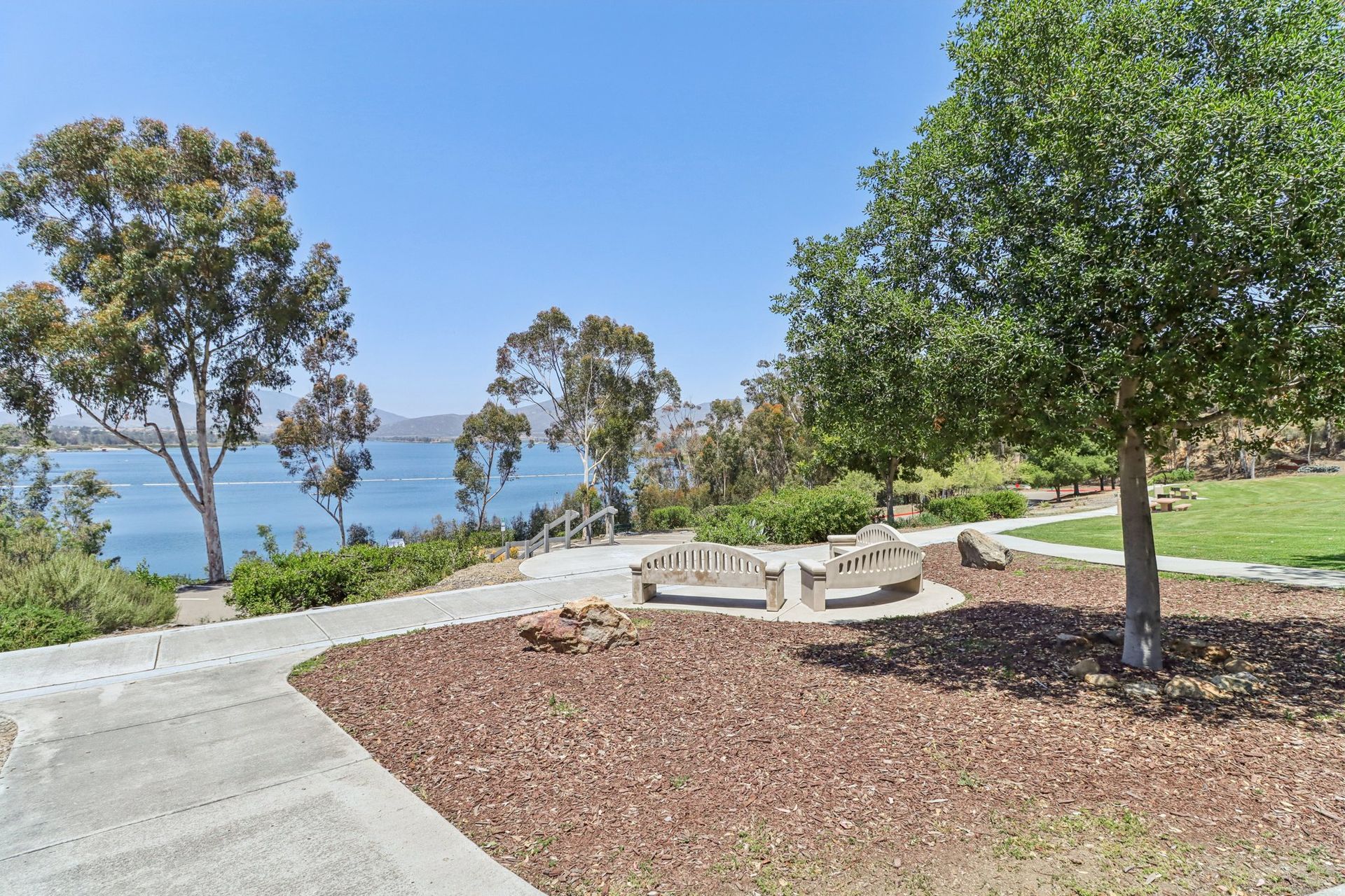 Park with a bench overlooking a blue body of water. Sunny day with trees and a path.