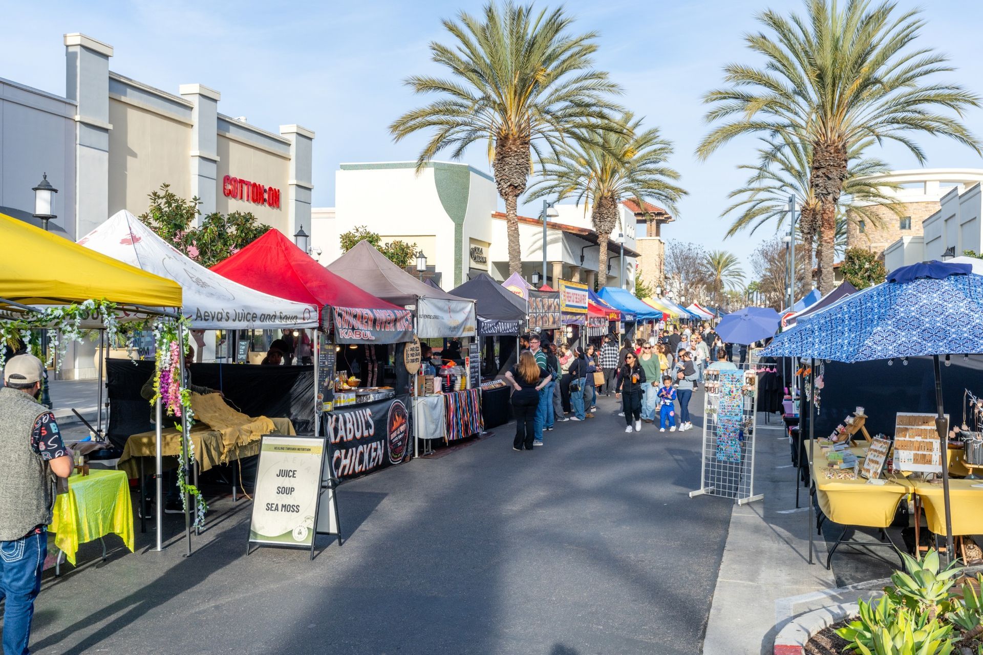 Street market with vendors under colorful tents; people browse and shop near palm trees.