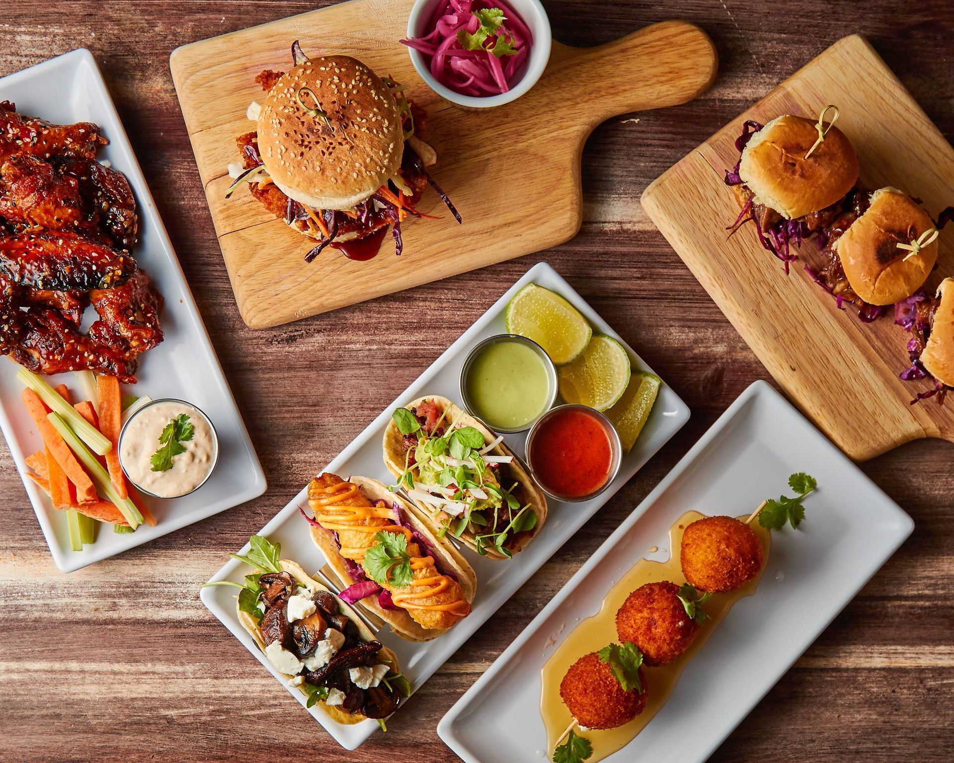 Assorted appetizers on wooden surfaces: wings, sliders, tacos, croquettes, and dips.