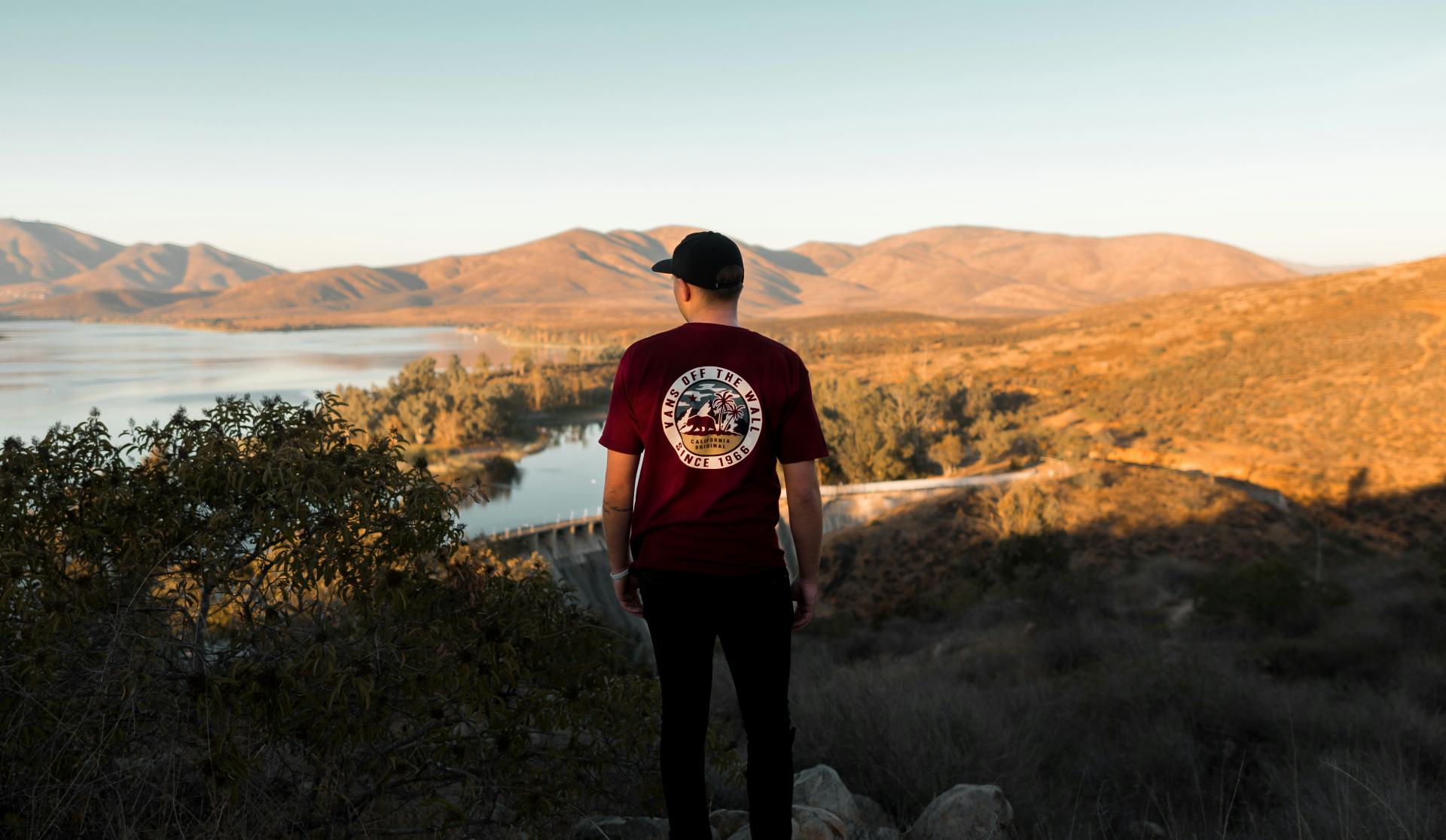 Man in maroon shirt, black cap, standing on a hill, looking at lake and mountains at sunset.