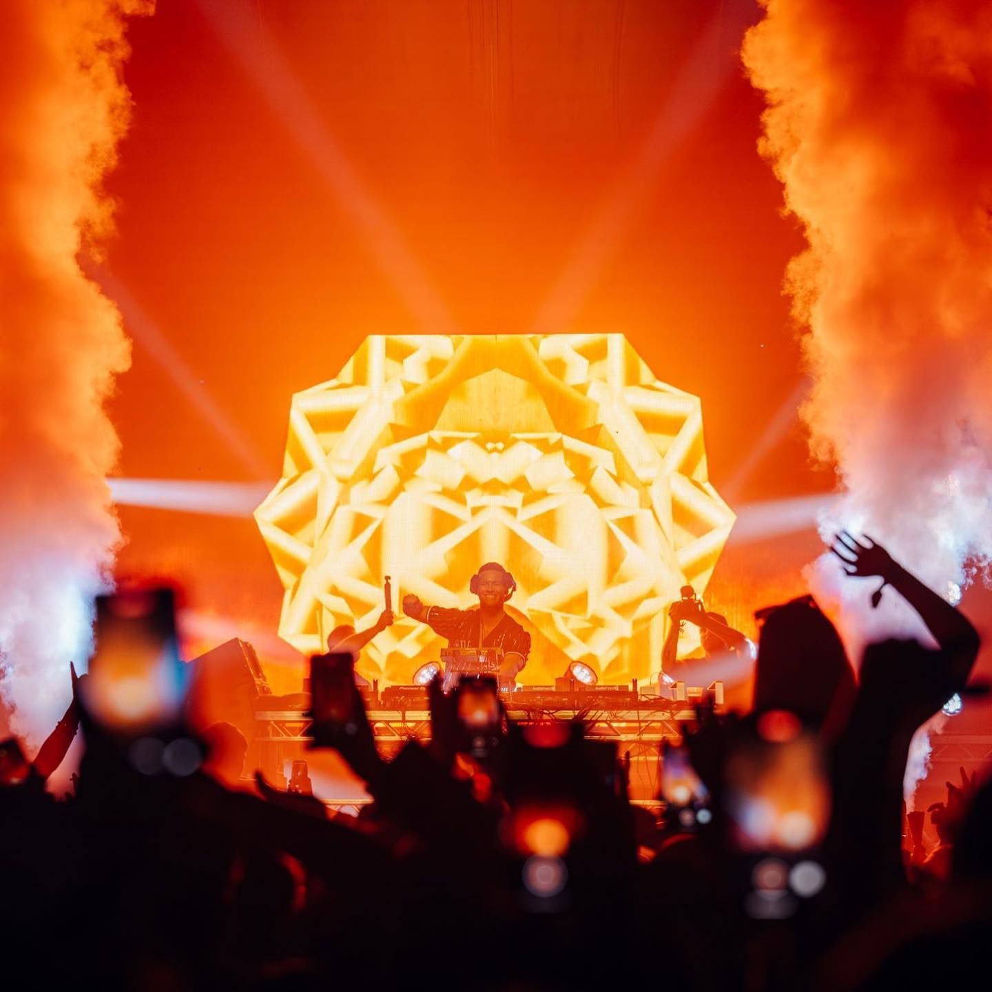 DJ performing on a bright orange-lit stage with crowd silhouettes and raised hands in the foreground