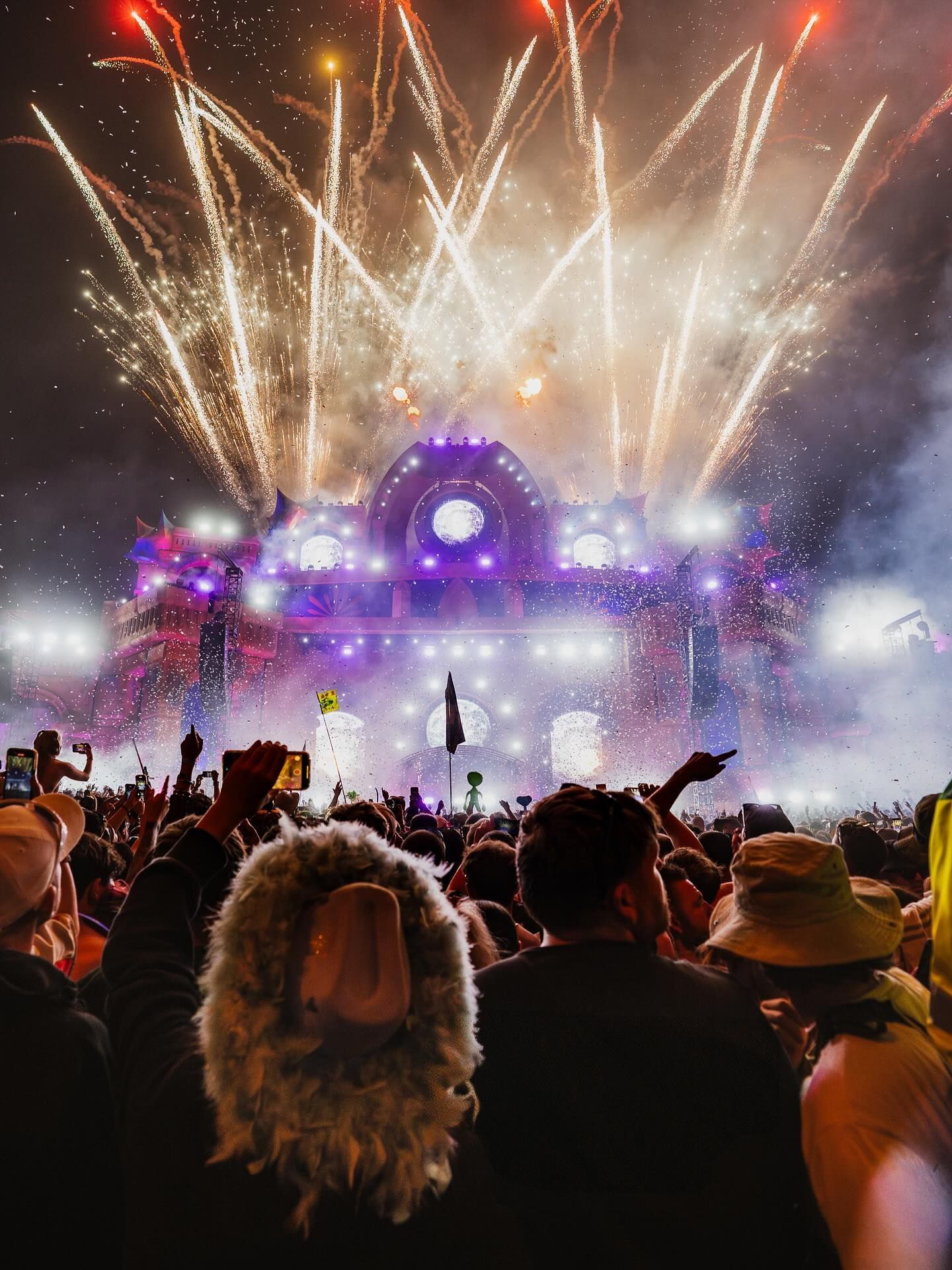 Crowd watching fireworks over a brightly lit festival stage at night