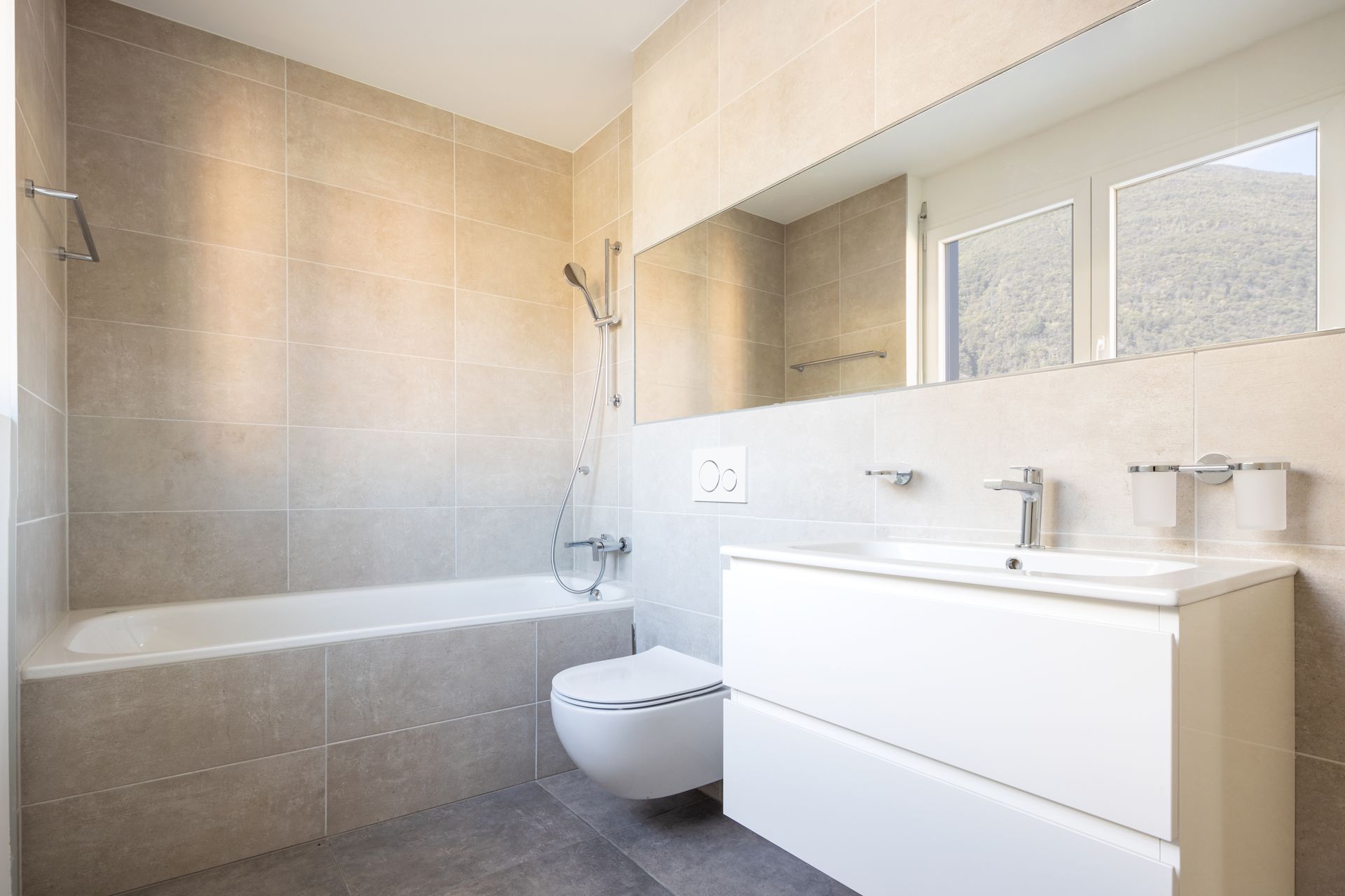 Modern bathroom with tiled bathtub, wall-mounted toilet, white vanity, and large mirror.