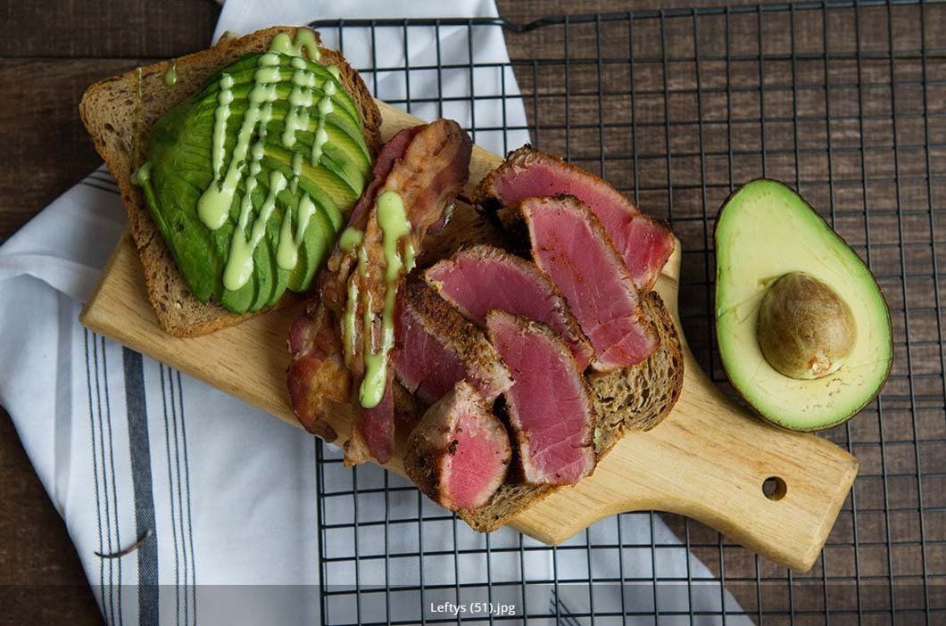 A wooden cutting board topped with meat , avocado and bacon.