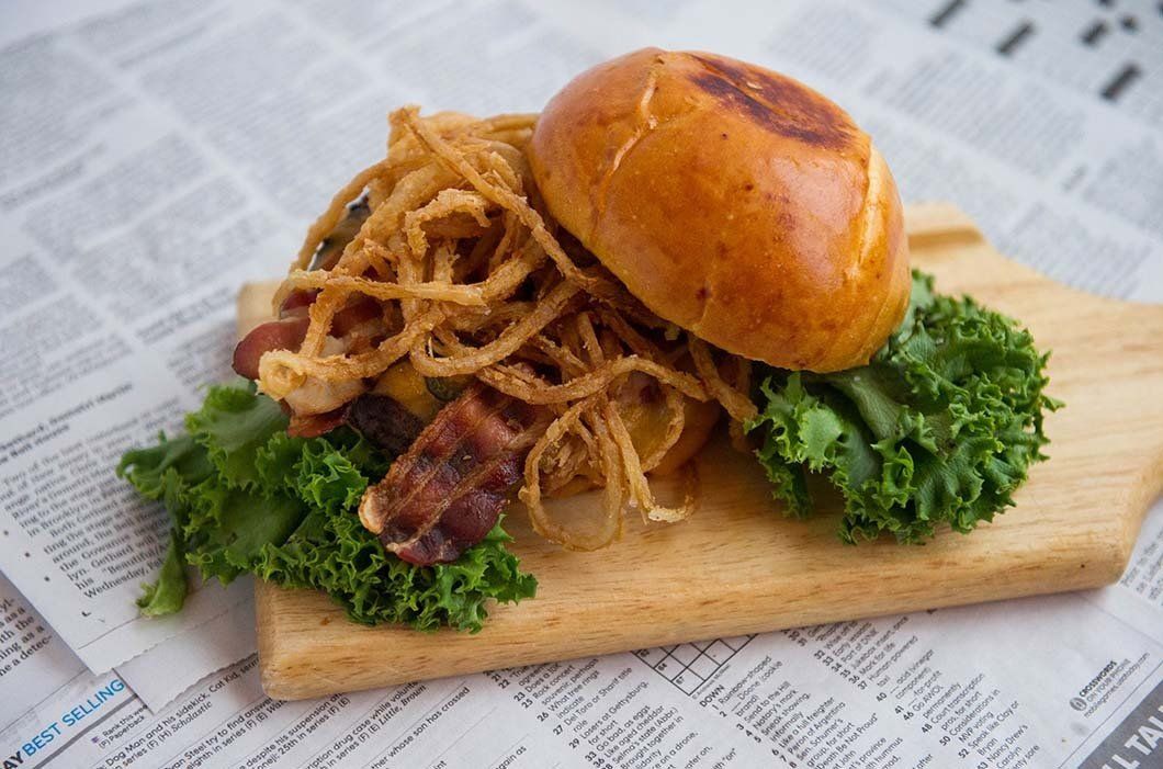 A hamburger with lettuce , bacon and onion rings on a wooden cutting board on top of a newspaper.
