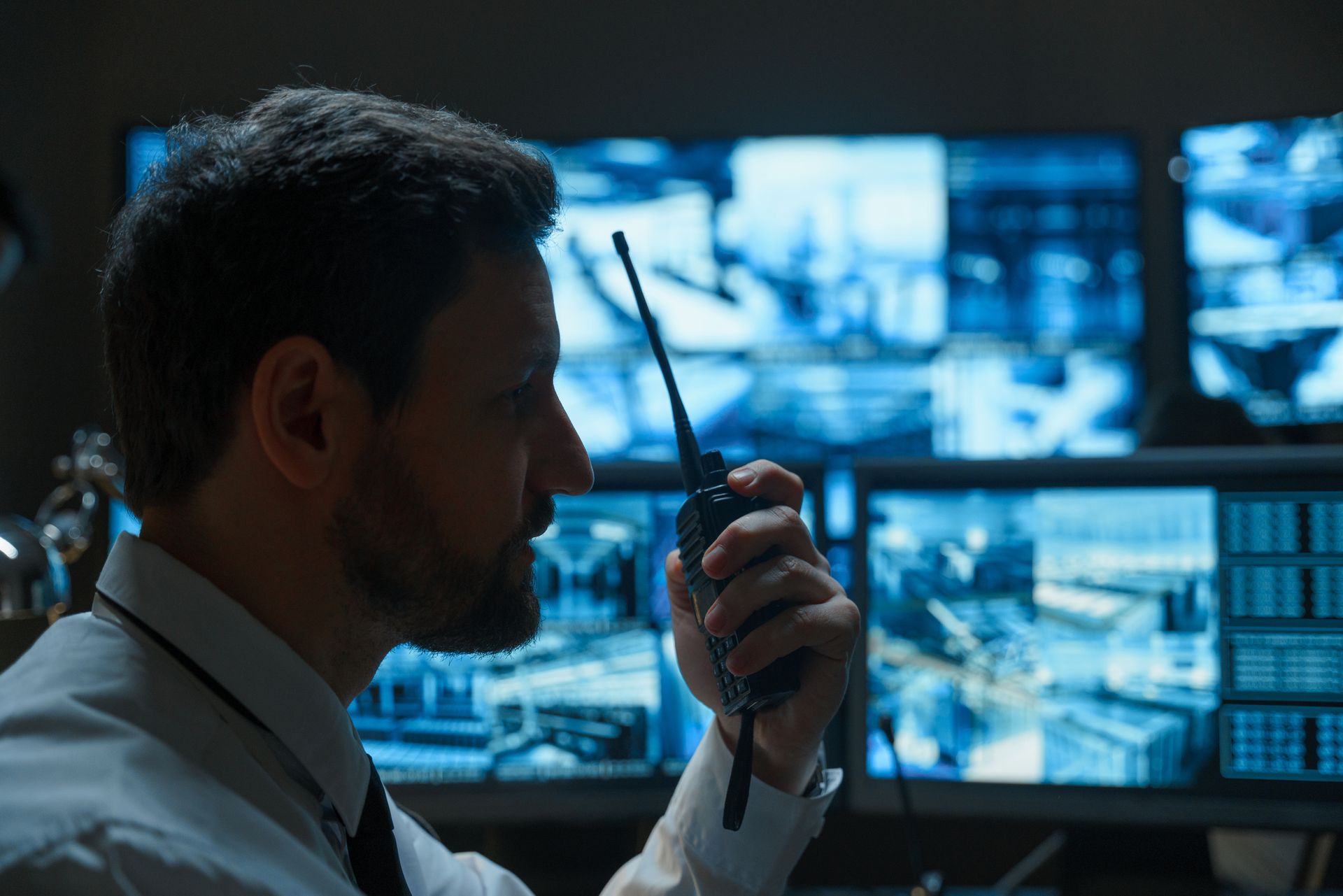 A man is talking on a walkie talkie in front of a bunch of monitors.