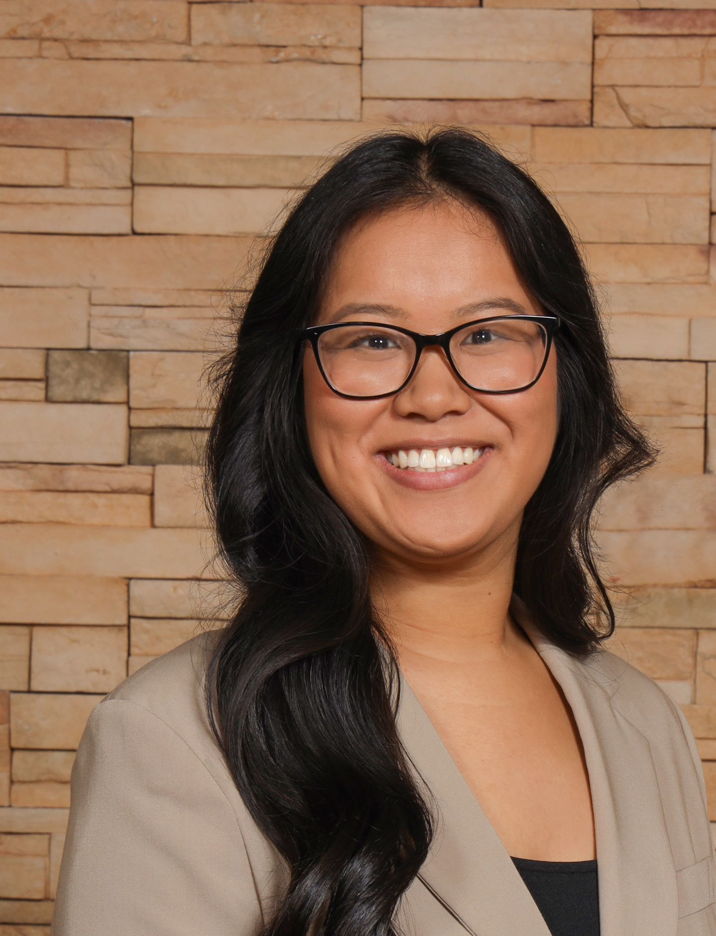 A woman wearing glasses and a tan jacket is smiling in front of a brick wall.