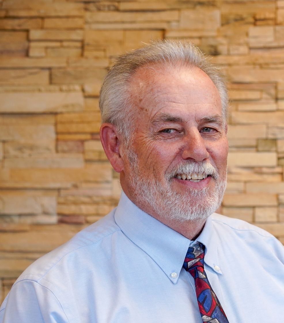 A man with a beard and tie is smiling in front of a brick wall.