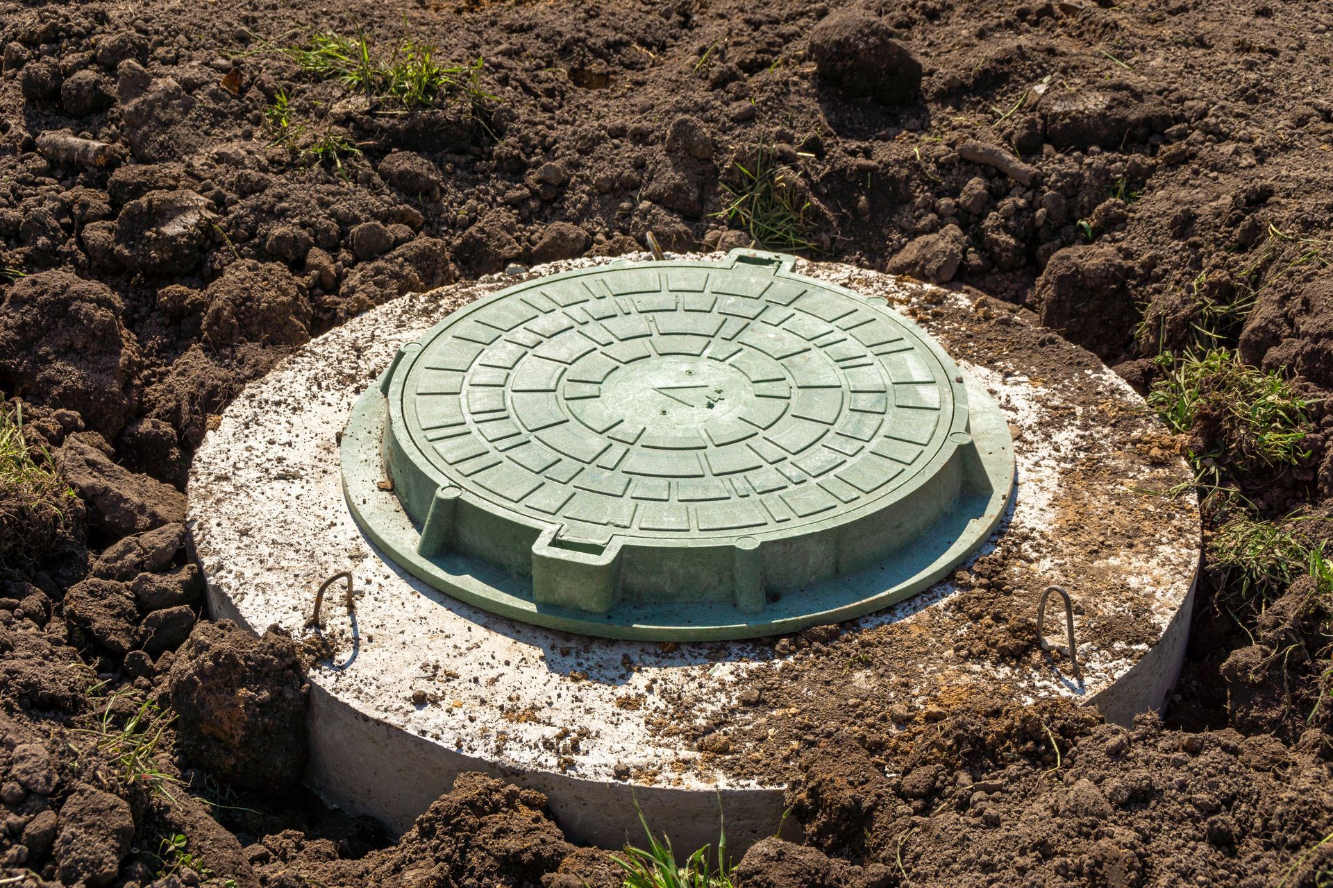 Green utility cover in dirt, set on a round concrete base with white stones around it