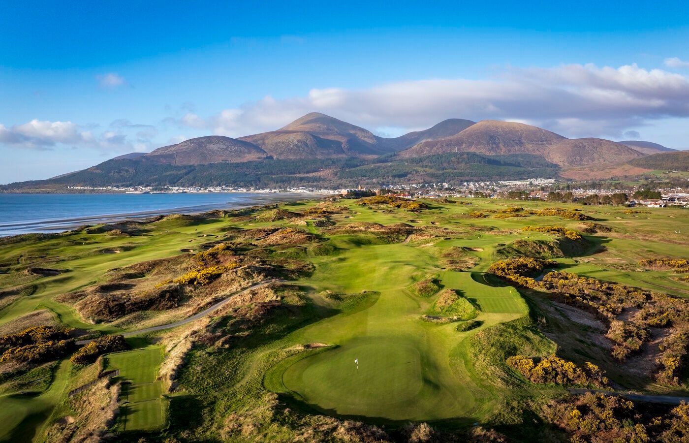 Golf course overlooking a coastal bay with mountains and a partly cloudy sky