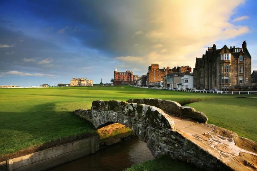 Stone bridge over a grassy field toward historic buildings under a dramatic sunset sky