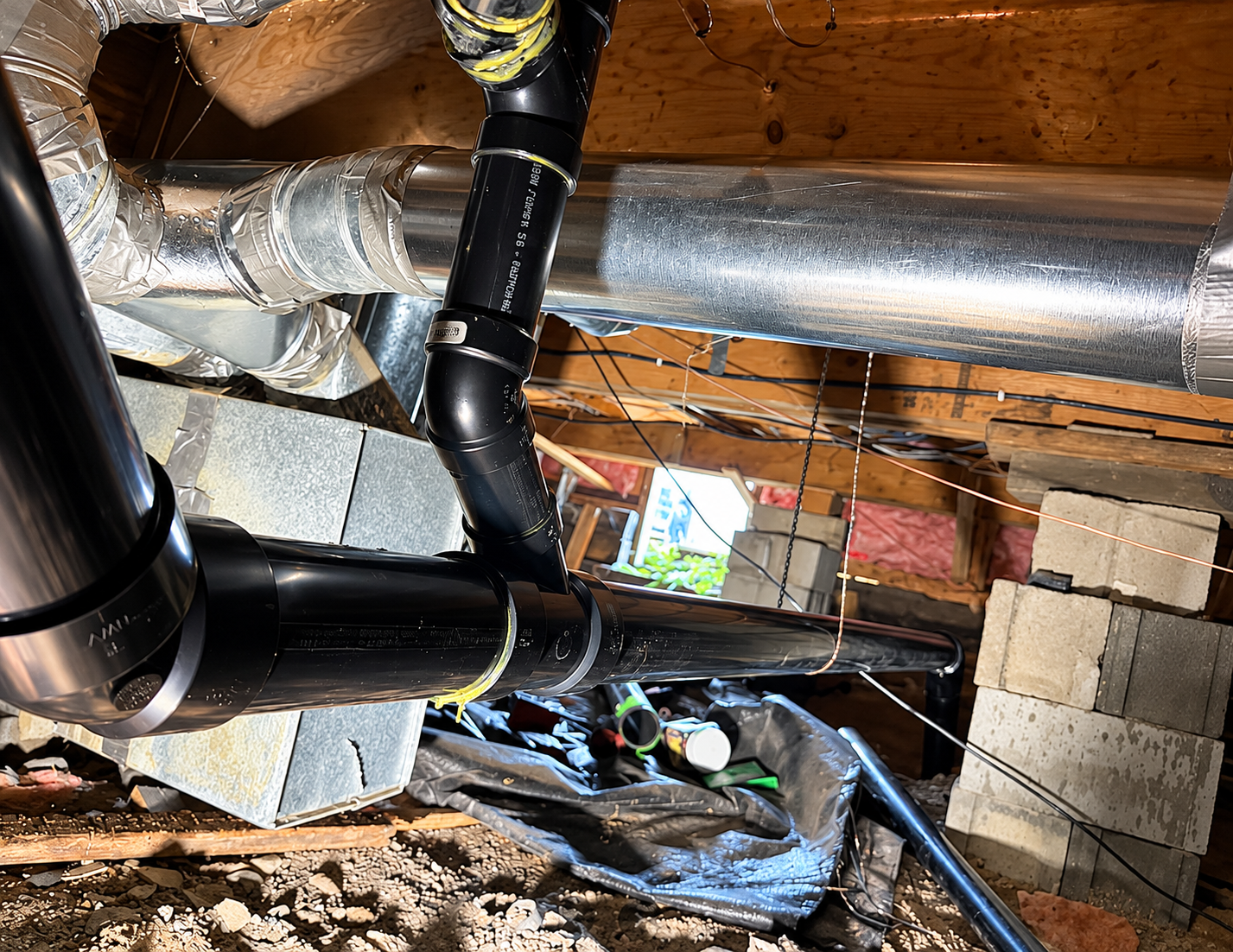 Insulation-damaged crawlspace with exposed pipes, wiring, and cinder blocks under a wooden floor.