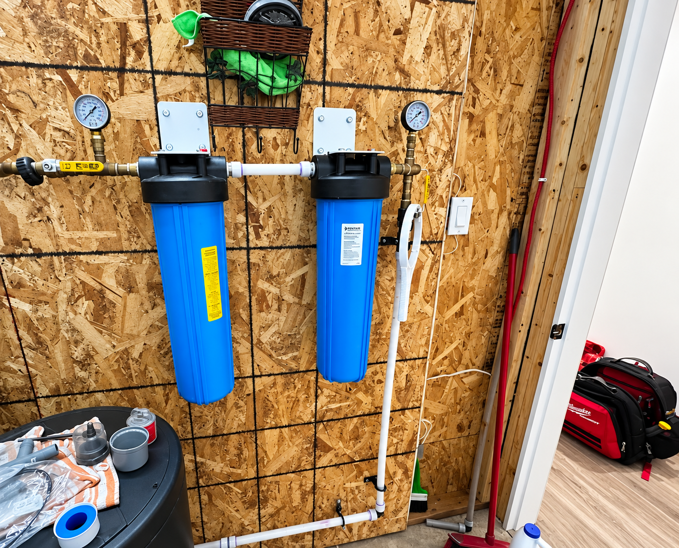 Two blue water filters mounted on an OSB wall in a utility area with gauges, pipes, and a small workbench.