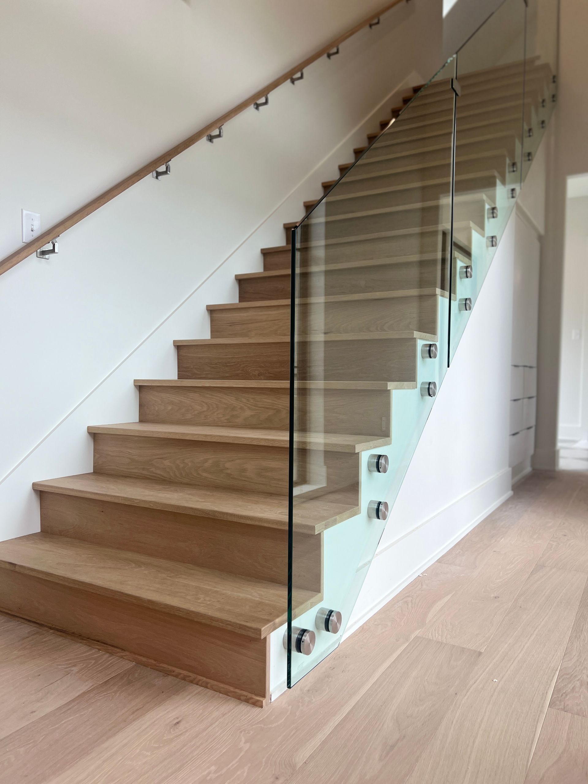 A staircase with light wood steps, a glass railing held by metal standoffs, and a wall-mounted wooden handrail.