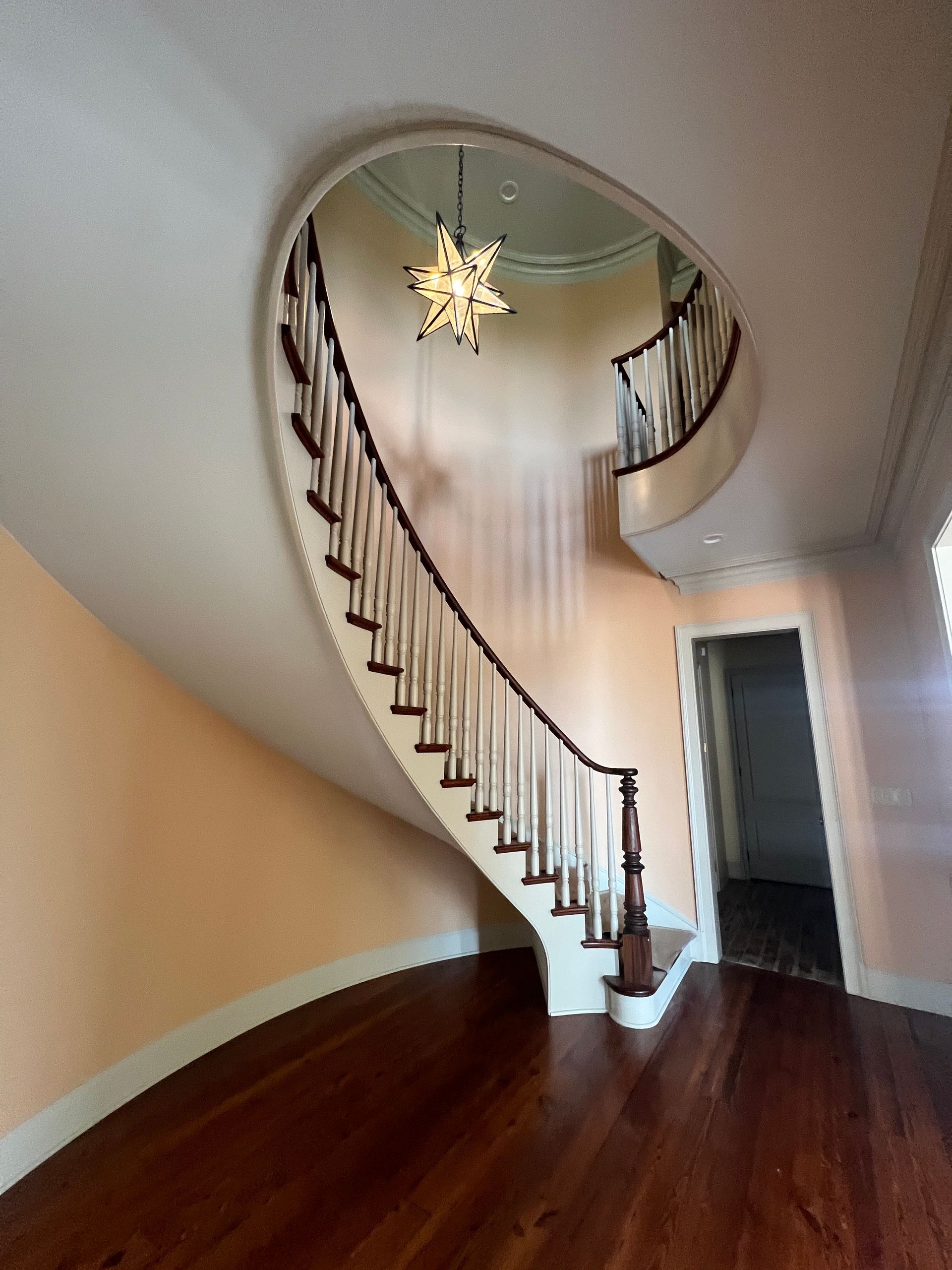 A curved wooden staircase with white railings rises in a spacious room with peach walls and dark hardwood floors.