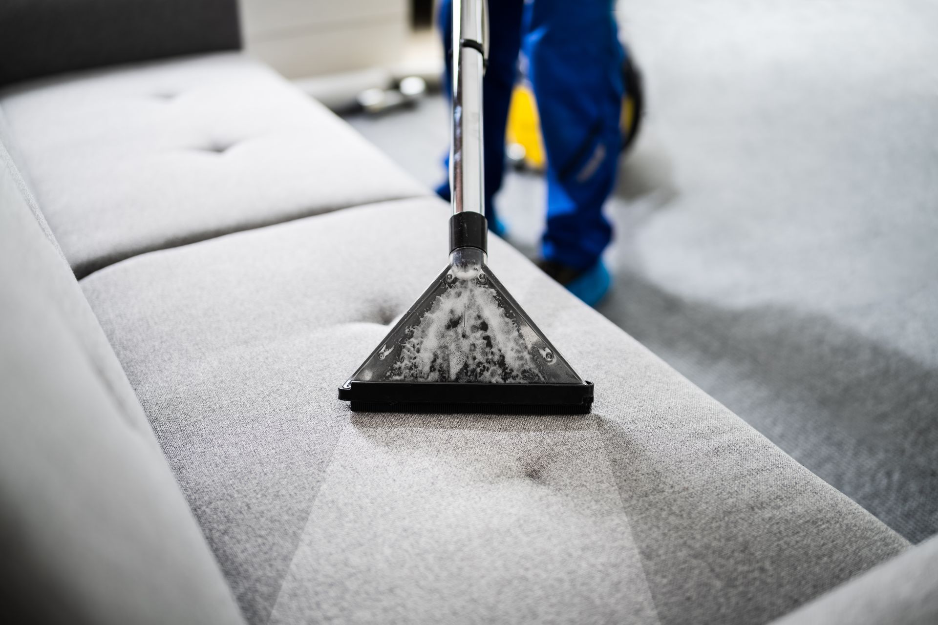 Une personne en salopette bleue nettoie un canapé gris avec un aspirateur.