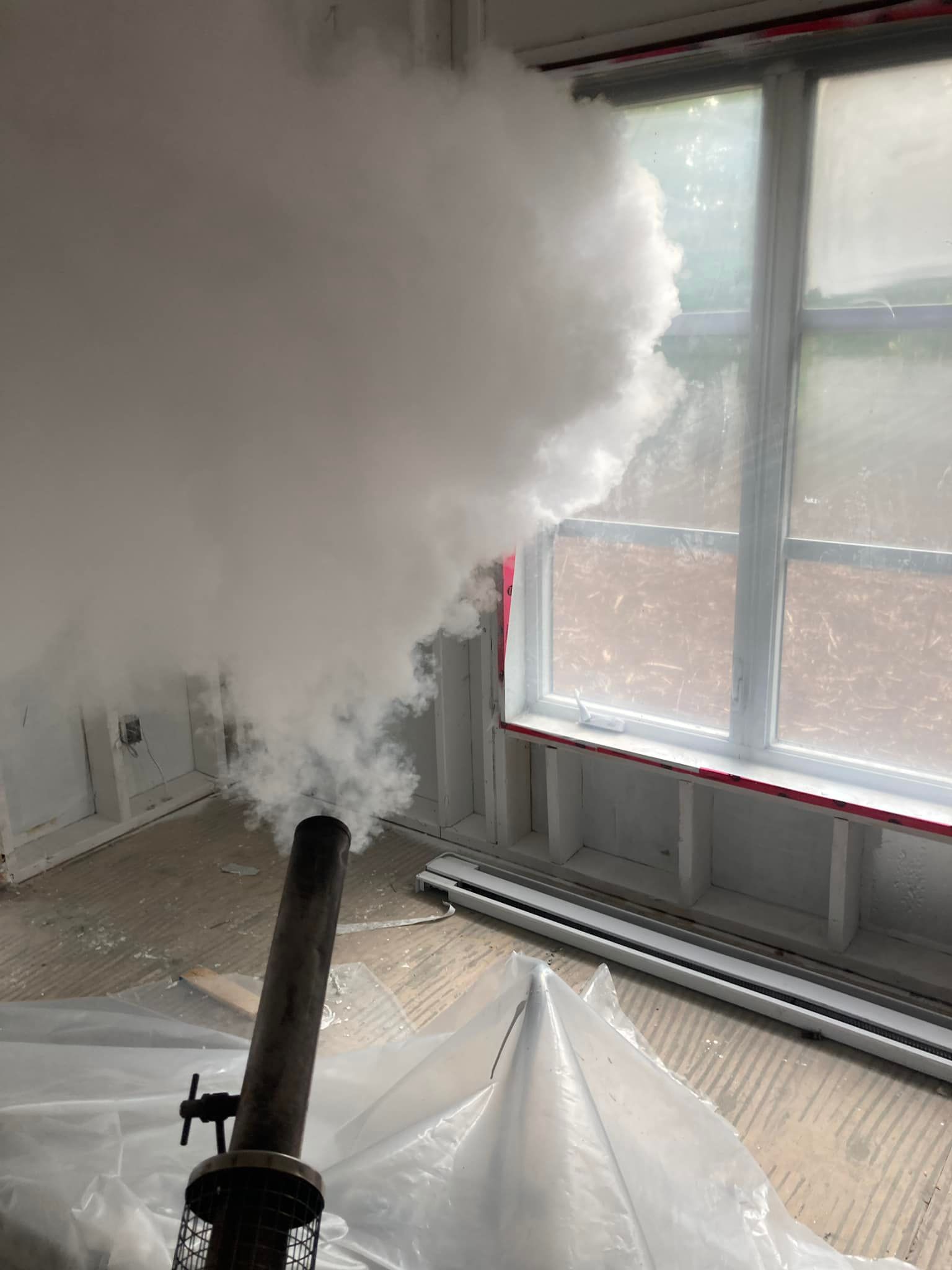 De la fumée s'échappe d'un appareil métallique et pénètre dans une pièce dotée d'une fenêtre ; une bâche en plastique transparent recouvre le sol.