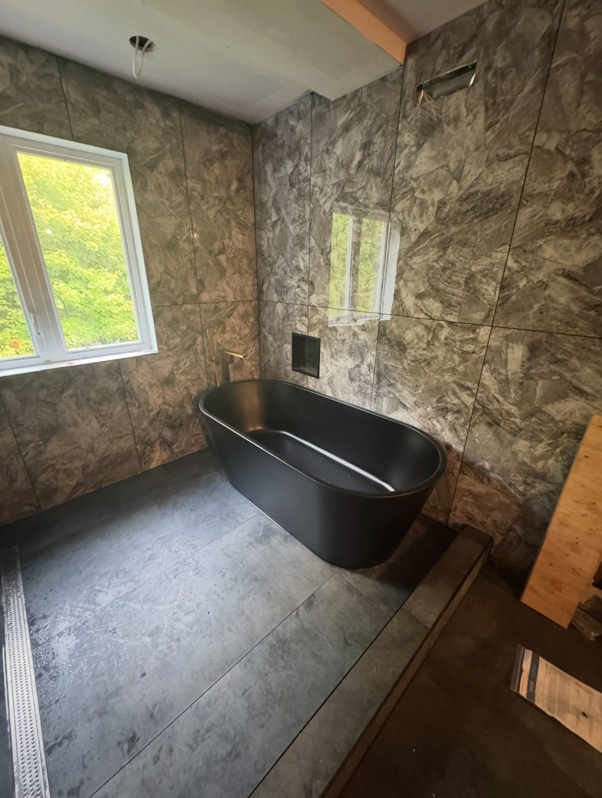 Salle de bains avec baignoire ovale noire, murs imitation marbre gris et fenêtre donnant sur la verdure.