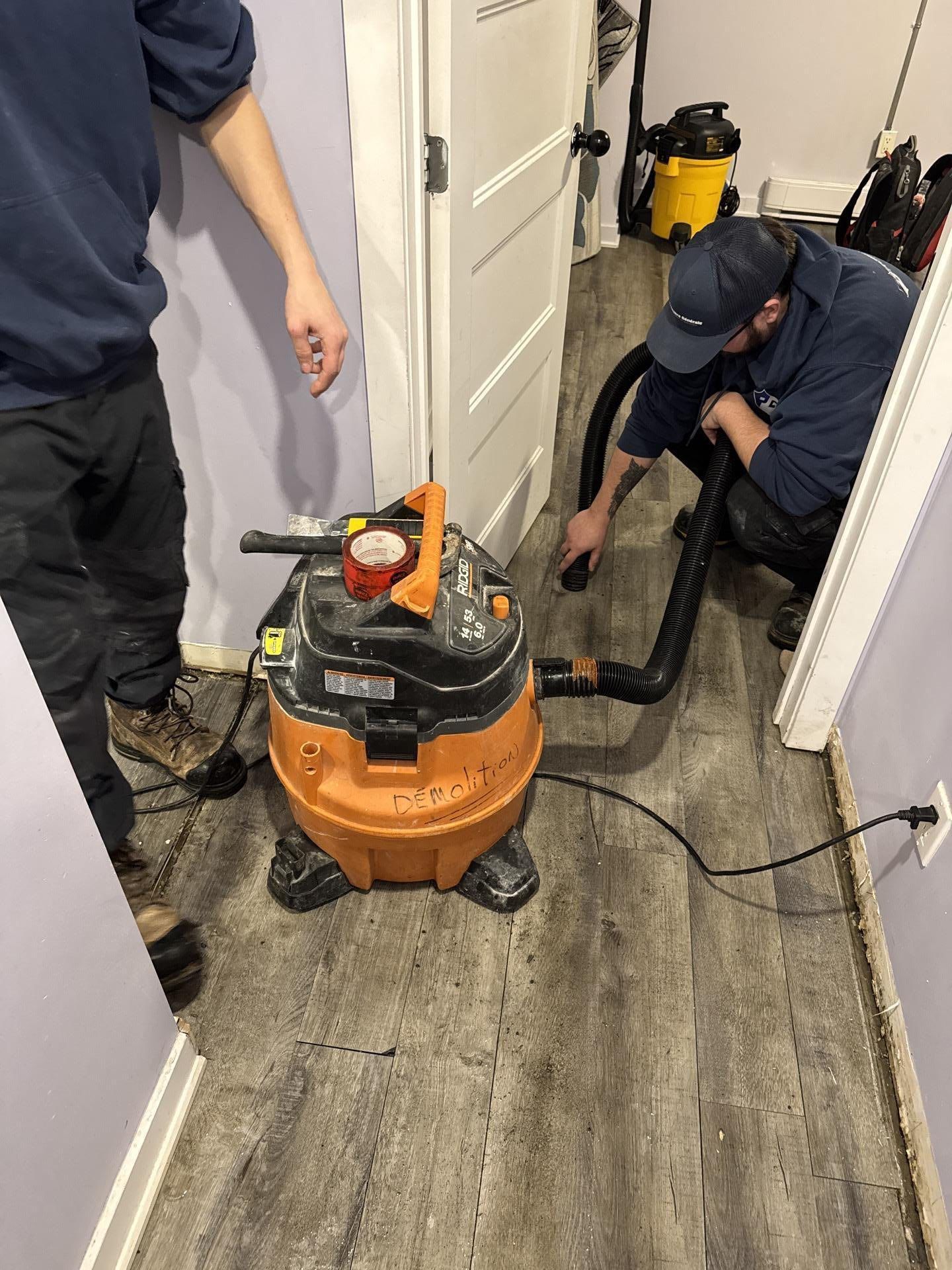 Deux personnes utilisent un aspirateur d'atelier dans un couloir au sol gris. L'une montre du doigt, l'autre actionne le tuyau.