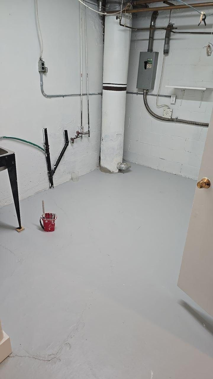 Sous-sol avec sol gris peint, plomberie, électricité et un tuyau cylindrique blanc. Une porte entrouverte se trouve à droite.