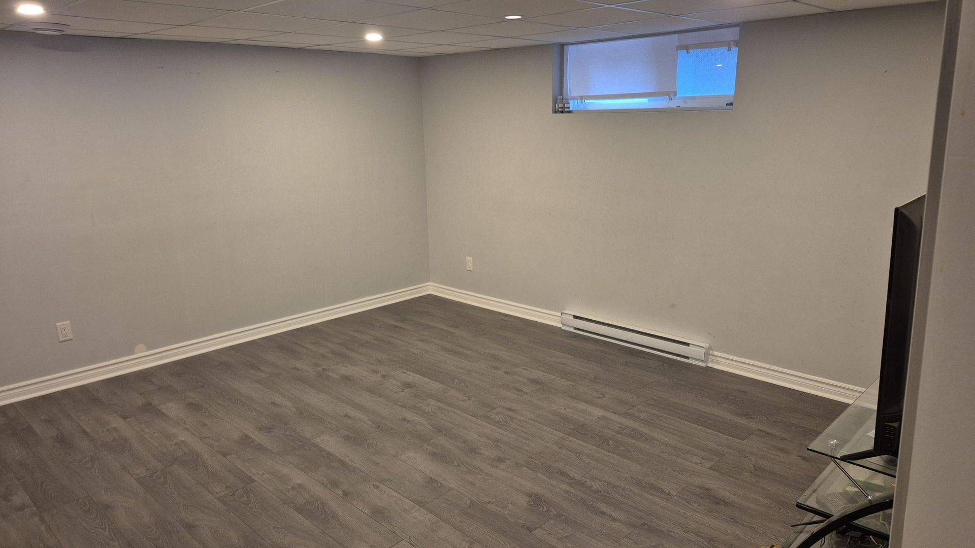 Pièce vide au sous-sol, murs gris et sol imitation bois. Petite fenêtre, radiateur plinthe et éclairage encastré.