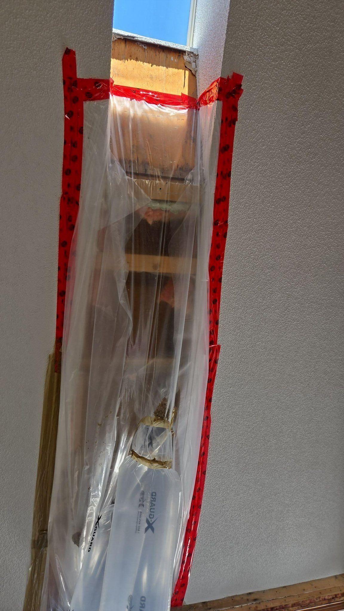 Une bâche en plastique transparent, fixée avec du ruban adhésif rouge, recouvre une étroite ouverture dans un mur blanc ; la lumière du jour est visible au-delà.