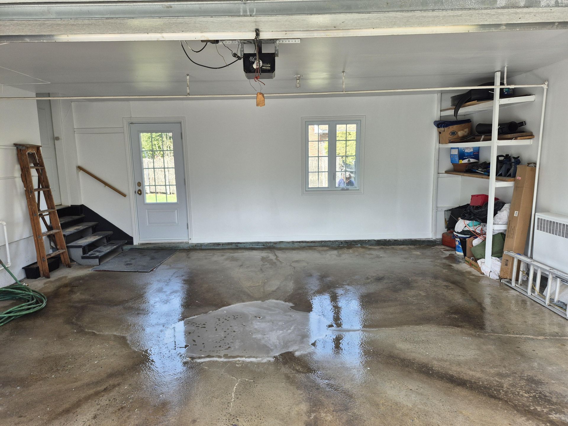Vue intérieure d'un garage au sol en béton mouillé. On aperçoit une porte et une fenêtre blanches, ainsi que des étagères et un escalier.
