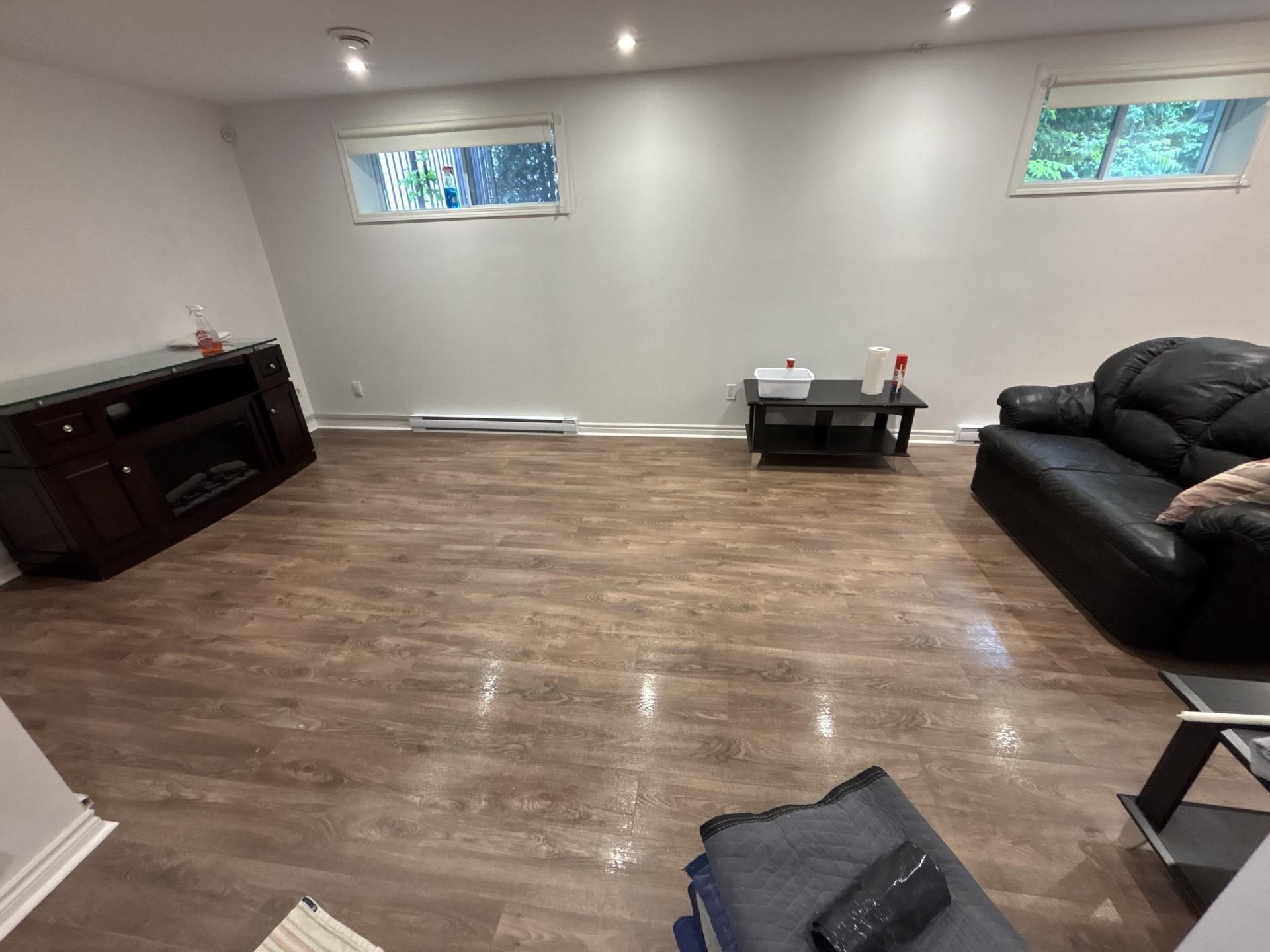 Salon avec parquet foncé, canapé en cuir noir, cheminée et petites fenêtres.