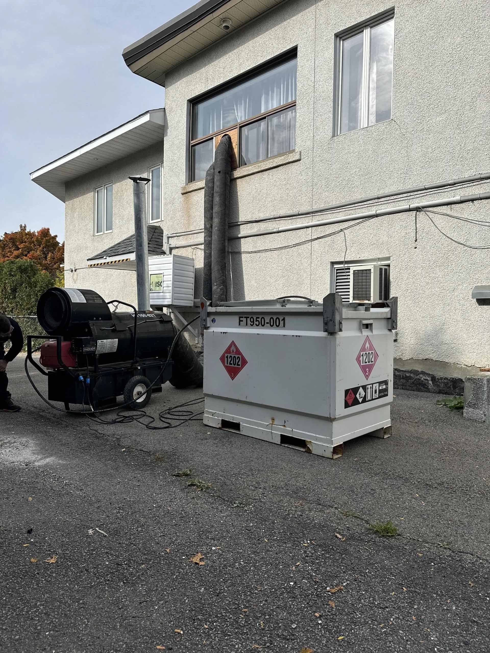 Bâtiment gris avec un système de chauffage et un conteneur à l'extérieur, reliés par un tuyau foncé.