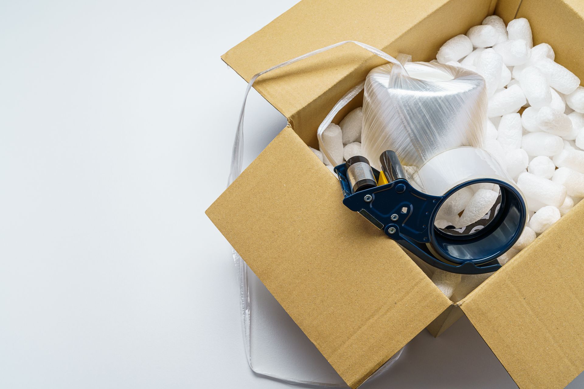 Open cardboard box with packing peanuts, bubble wrap, and a black tape dispenser on a white surface