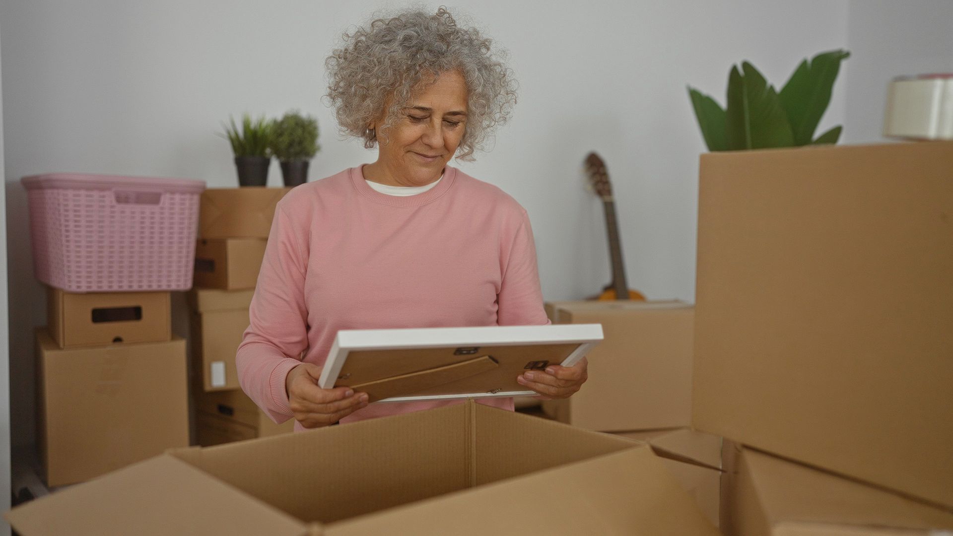 Person unpacking framed art from a cardboard box in a living room with moving boxes and plants.