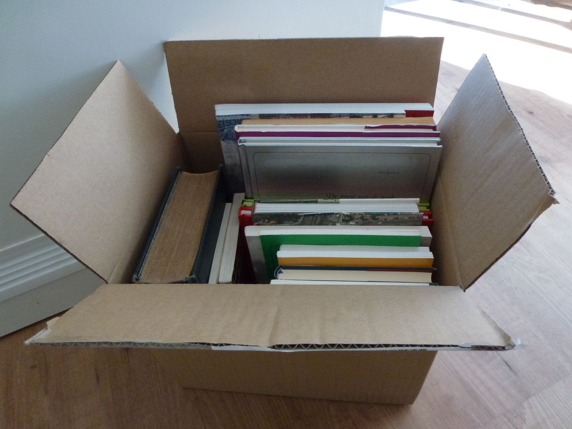 Open cardboard box filled with assorted books and papers on a wooden surface.