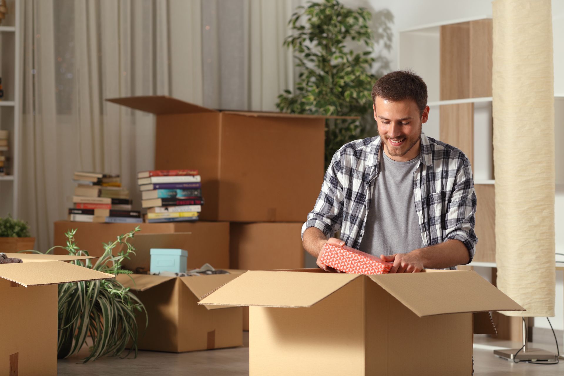 Man packing boxes in a bright room surrounded by stacked moving boxes and plants
