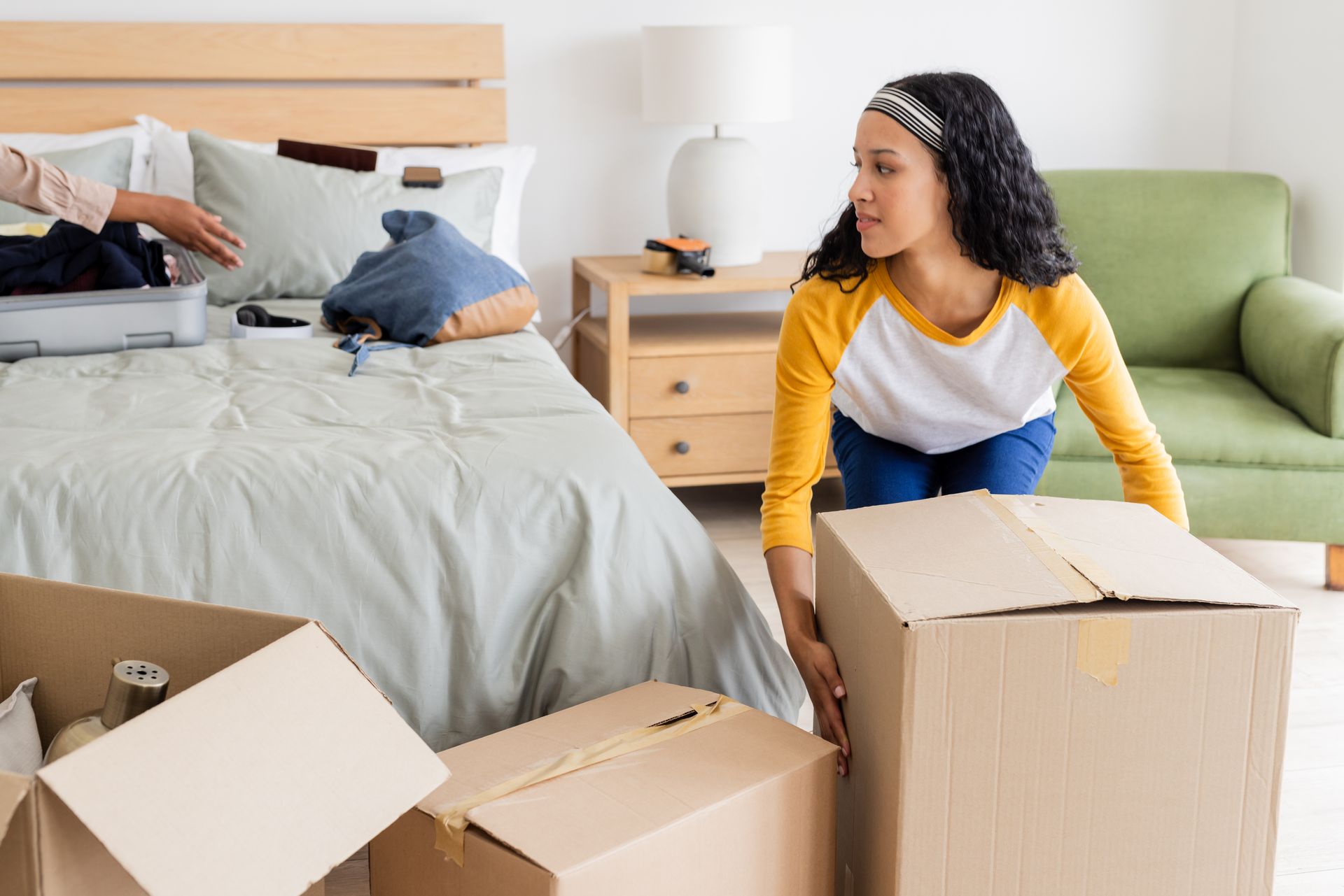 Person kneeling by moving boxes in a bedroom, with another person arranging pillows on the bed