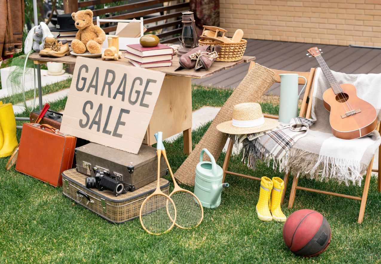 Garage sale table with toys, baskets, hats, boots, a guitar, and a sign on a grassy lawn