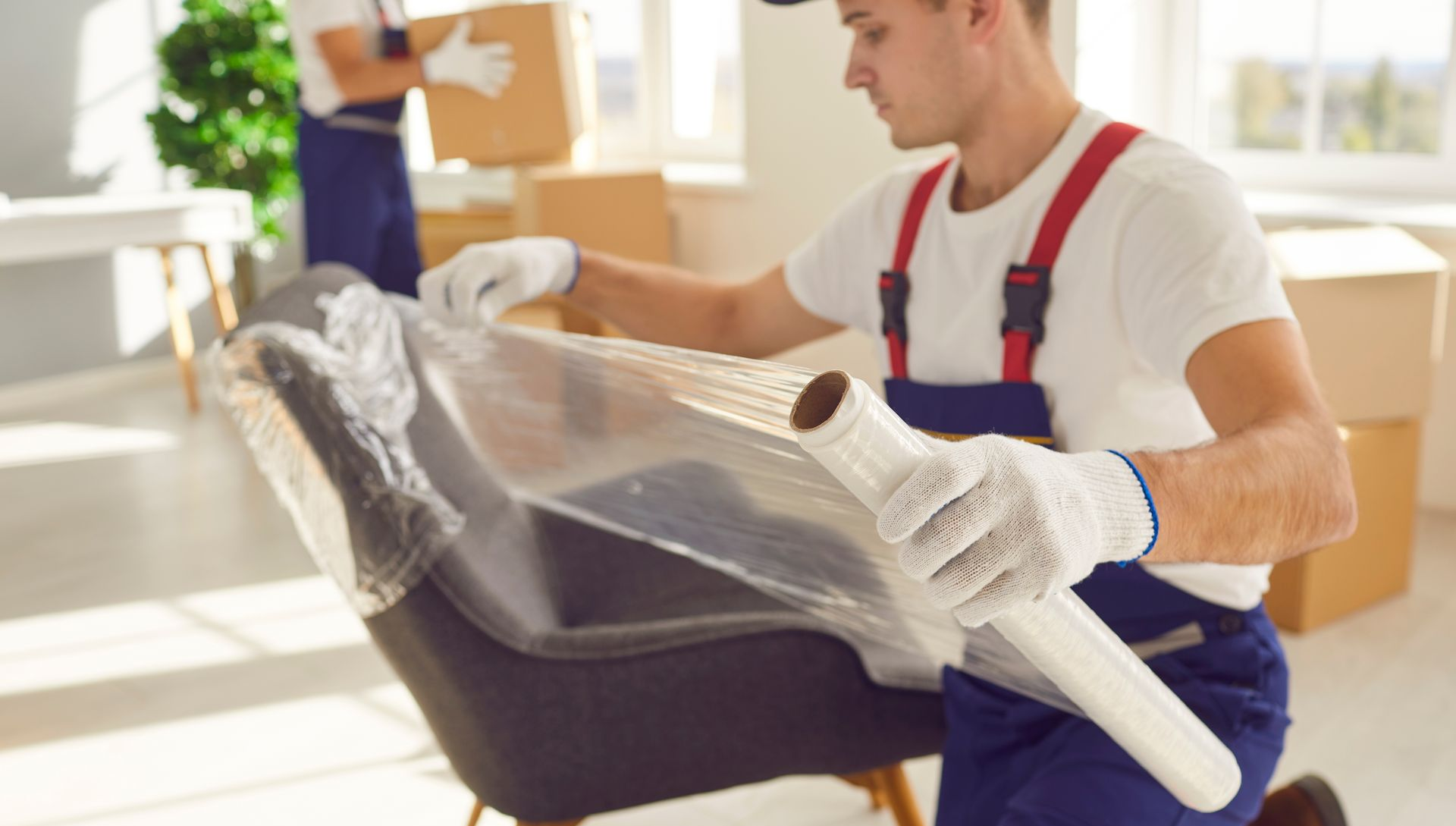 Man in overalls wrapping a chair with plastic for moving; another person in the background carries a box.