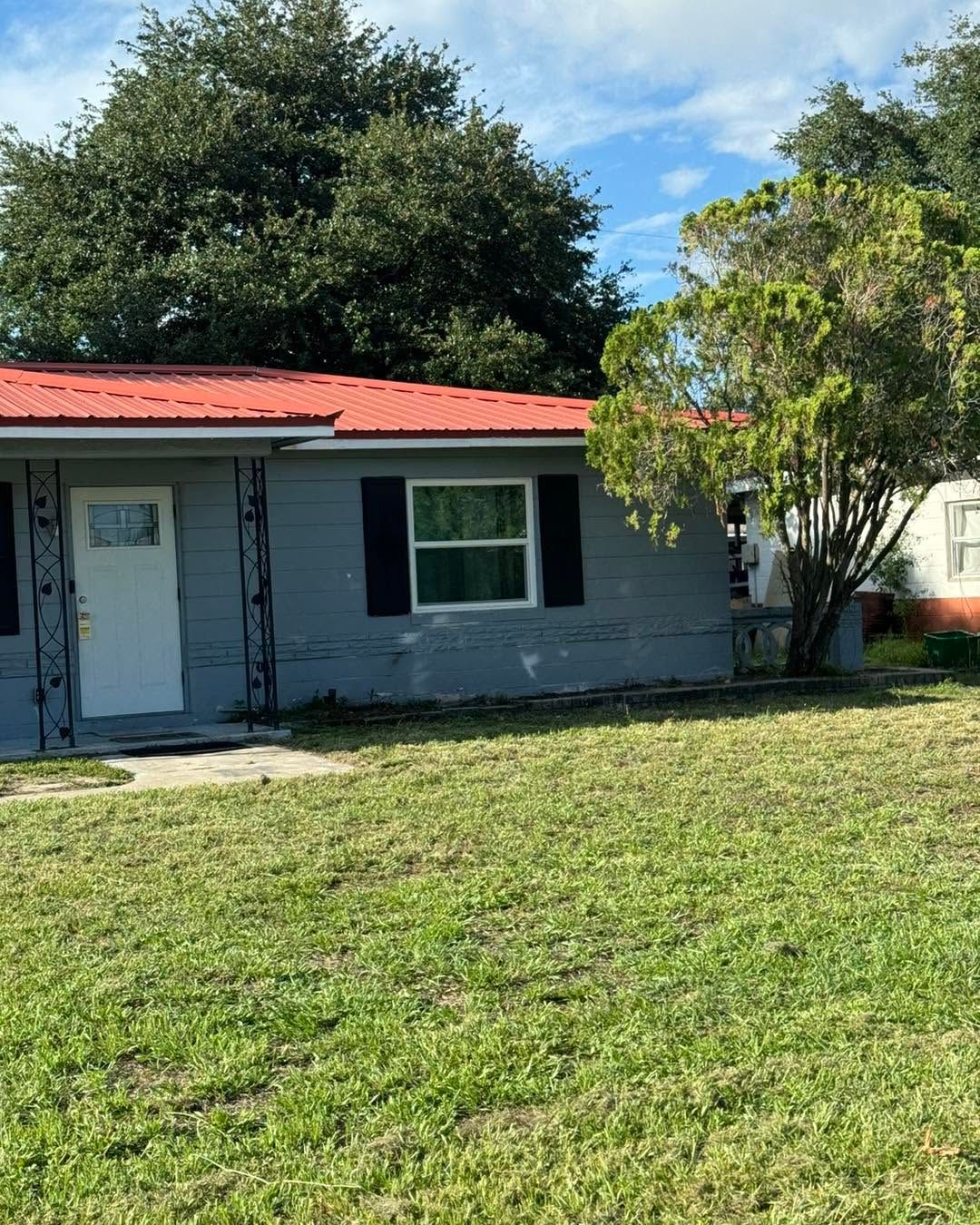 Single-story gray house with a red roof and front yard under trees on a sunny day