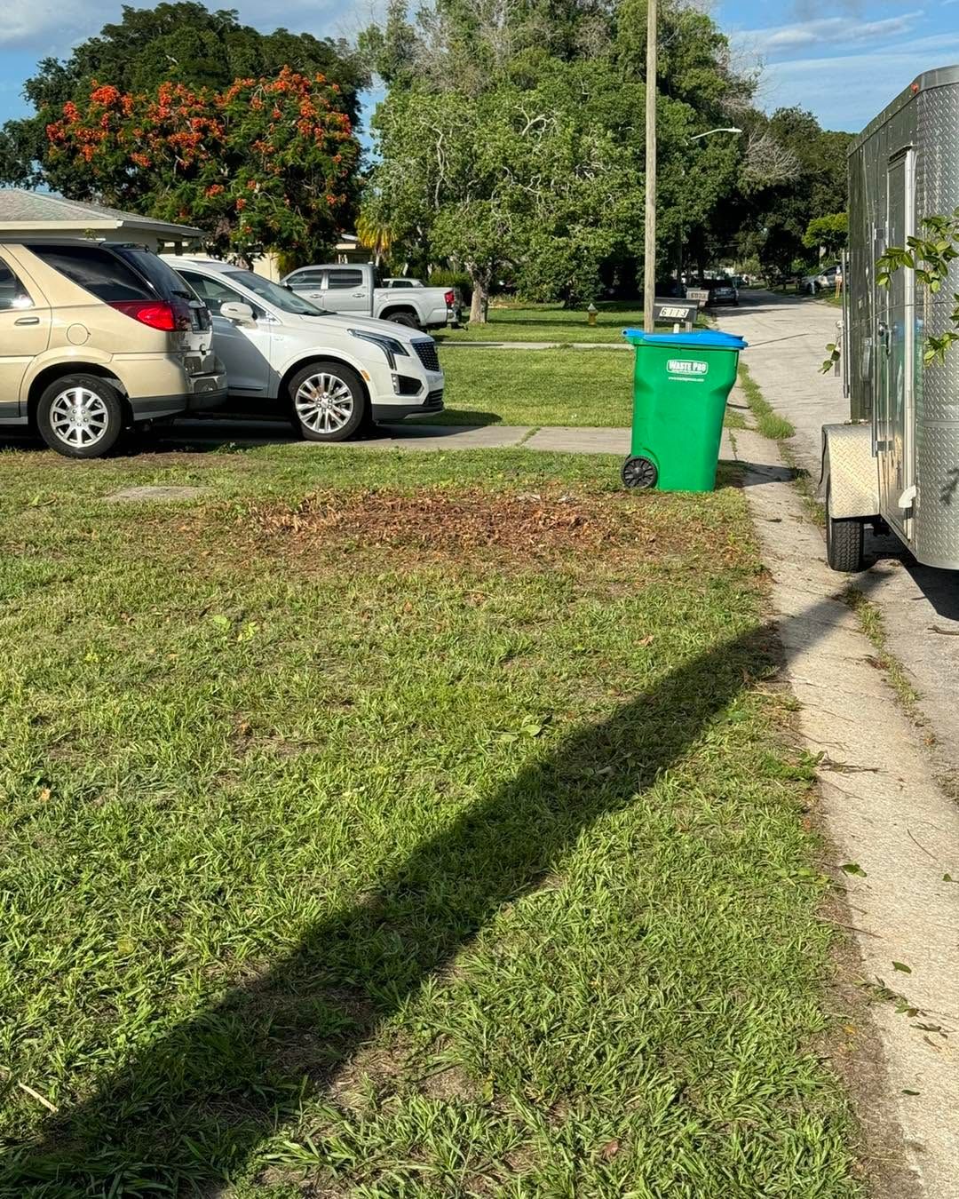 Parked cars beside a grassy roadside with a green trash bin and a truck on the right.