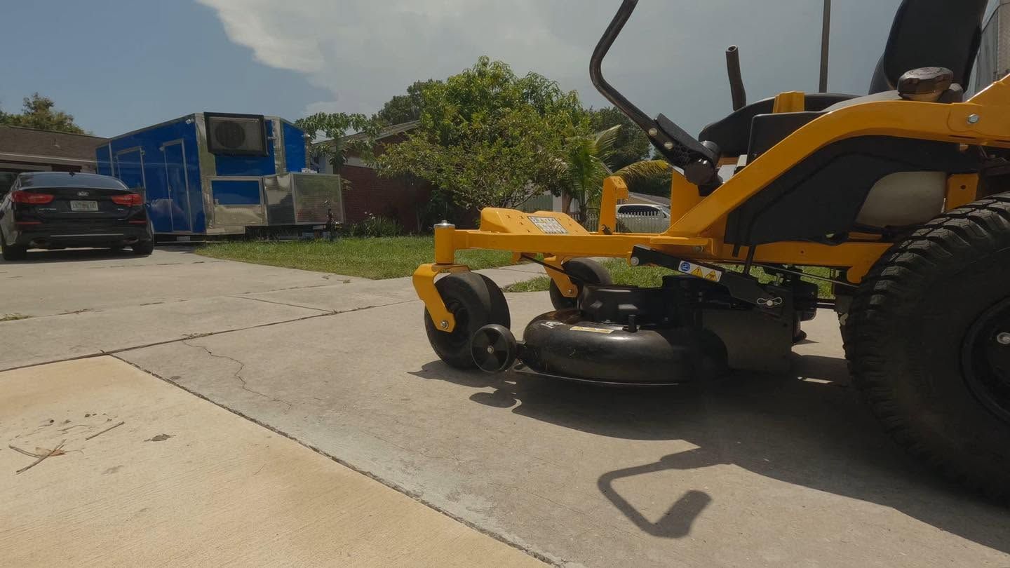 Yellow lawn mower on a concrete road near a blue truck under a cloudy sky