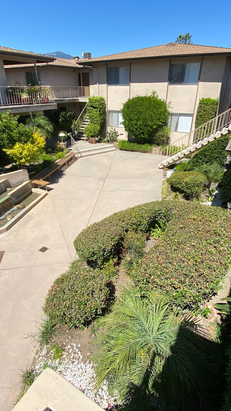 Two-story beige apartment buildings with a concrete courtyard, stairs, and manicured green landscaping.