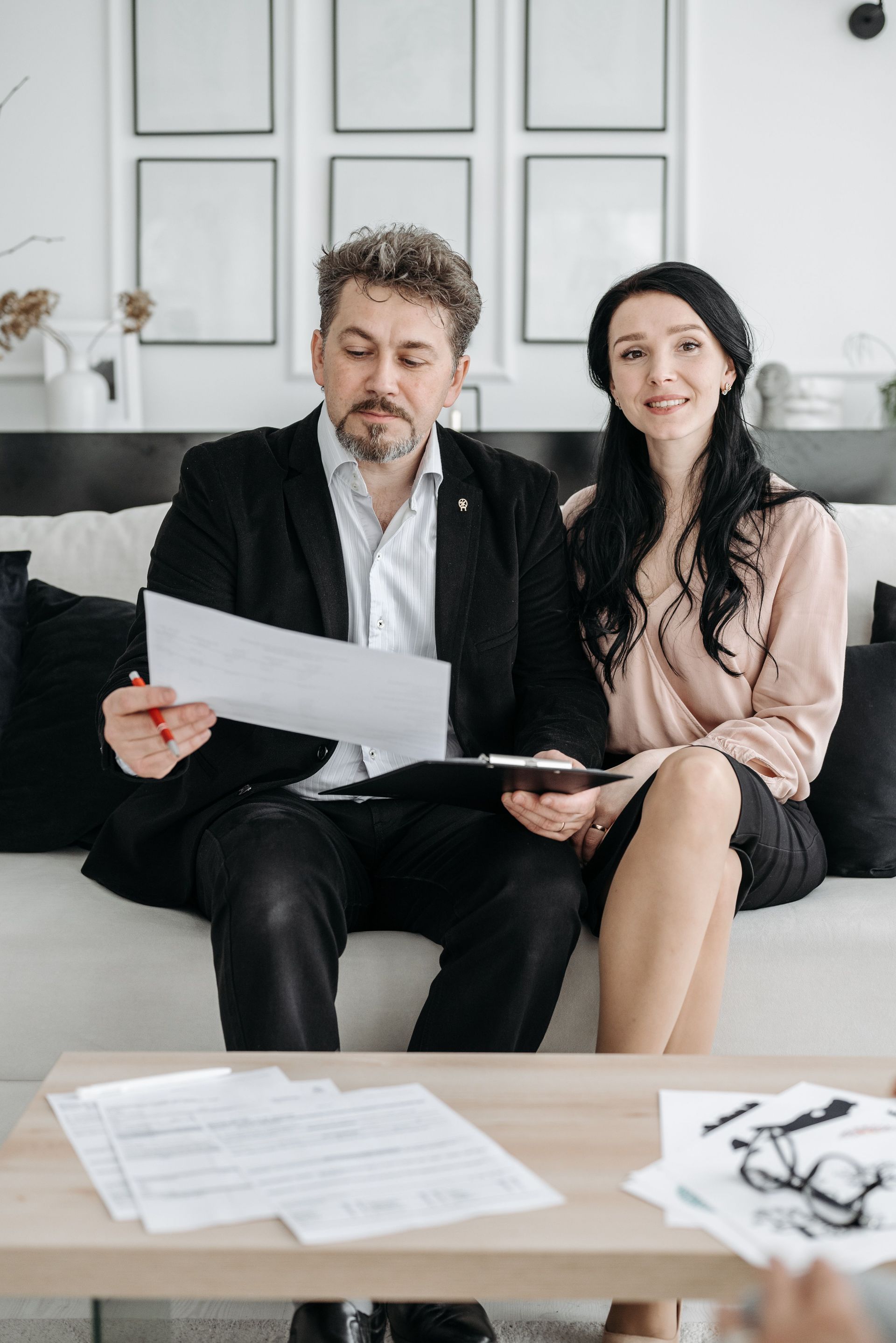 Two people reviewing paperwork on a couch in a bright living room with documents spread on the table